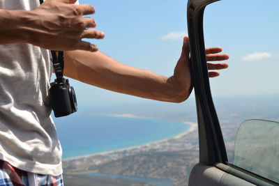 Midsection of man with camera holding car door against sky