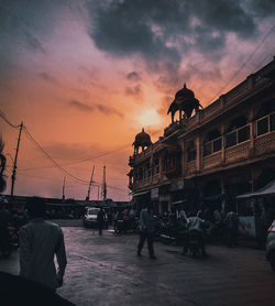 People on street by buildings against sky at sunset