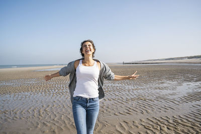 Cheerful young woman with arms outstretched walking at beach against clear sky