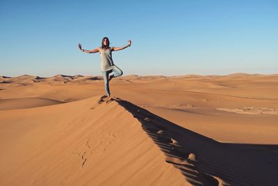 Woman practicing yoga on sand dune at desert against clear blue sky