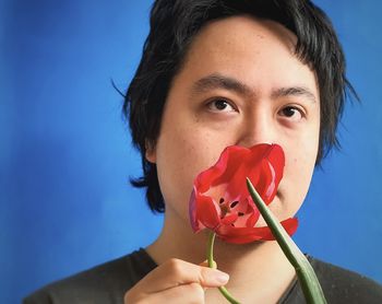 Close-up portrait of a young man holding red tulip against mouth against blue background.