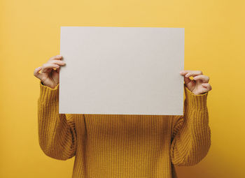 Woman in an orange sweater holds a blank sheet of paper on a yellow background. place for an 