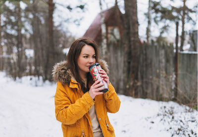 Young brunette woman in yellow jacket with thermo mug at cottage village in winter forest
