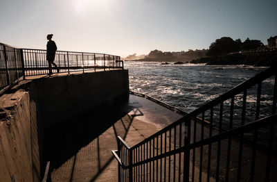 Woman walking by railing and sea against sky during sunset