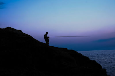 Silhouette man fishing on rock against clear sky at dusk