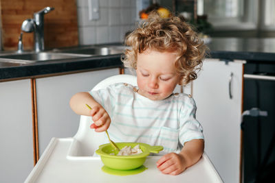 Caucasian curly kid boy sitting in high chair eating cereal puree with spoon. healthy eating 