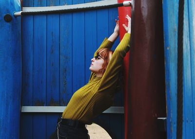 Side view of woman with arms raised standing by wall outdoors