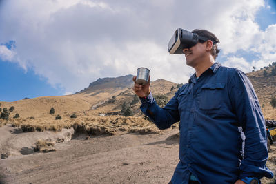 Man holding mug while wearing virtual reality headset against mountain