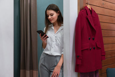 Woman using mobile phone while standing by coat at home