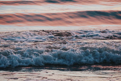 Waves rushing on shore against sky during sunset