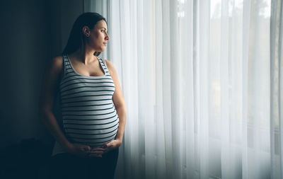 Young woman looking away while standing against window