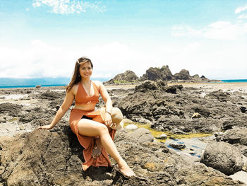 Young woman sitting on rock at beach against sky