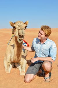 Smiling man looking at camel while kneeling on desert