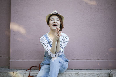 Portrait of smiling young woman sitting against wall outdoors