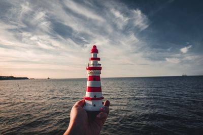 Close-up of person holding figurine lighthouse by sea against sky