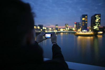 Woman photographing illuminated skyscrapers by river from camera