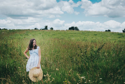 Teen girl standing in a grassy field holding a sun hat on a windy day.