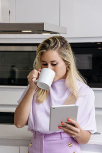 Female standing in kitchen and drinking fresh morning coffee while reading news on tablet in morning and having breakfast