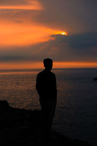 Rear view of man standing at beach against sky during sunset