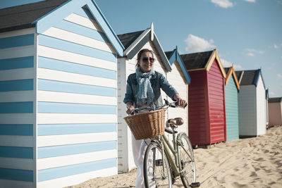 Woman walking with bicycle on sand by beach huts on sunny day