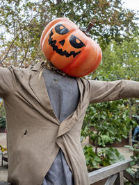 Scarecrow in ragged clothing with a classic pumpkin head, on a stake in front of tree leaves.