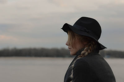 Young woman wearing hat at lakeshore against sky during sunset