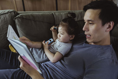 Father with male toddler reading book while lying on sofa in living room