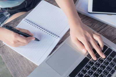 Cropped image of woman writing on book while using laptop at table