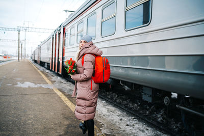 Girl on the platform of the station with a red backpack and a bouquet of tulip flowers