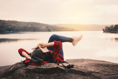 Full length of young woman sitting on rock by lake against sky