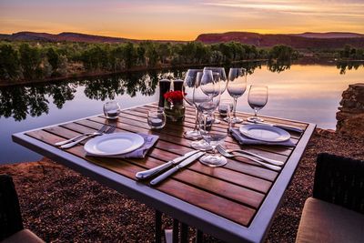 Chairs and table by lake against sky during sunset