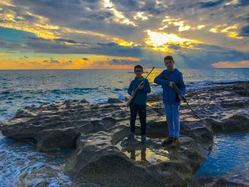 Men standing on rock by sea against sky during sunset