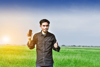 Portrait of man holding mobile phone while standing on grassy field against sky