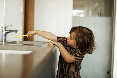 Boy holding toothbrush under running water from faucet