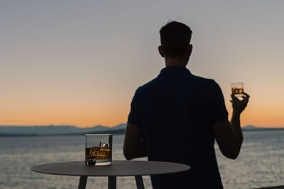 Rear view of man holding whiskey glass by sea during sunset