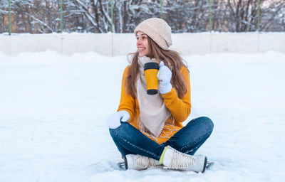 Portrait of happy young woman with ice skates sitting on the snow and holding mug with coffee
