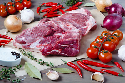 Close-up of large piece of fresh pork meat on kitchen table surrounded by fresh vegetables. 