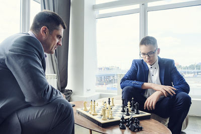 Dad and son are sitting at a table by a wide window and playing chess making moves with pieces