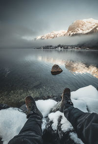 Low section of person on snowcapped mountain by lake against sky