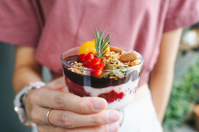 Close-up of woman holding granola and yogurt with fruits in glass bowl on table