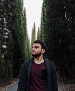 Young man looking away while standing against trees