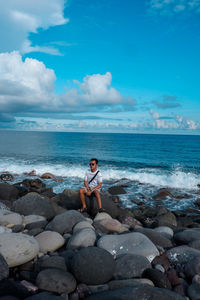 Rear view of man sitting on rock at beach against sky