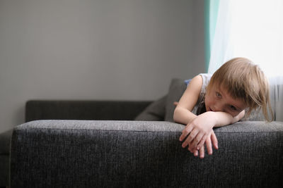 European toddler child sits and gets bored on gray sofa in bright living room, minimalist interior