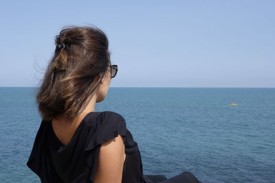Young woman wearing sunglasses sitting at beach against clear sky