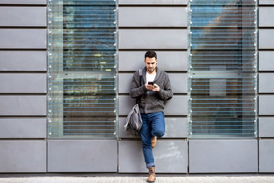 Front view of a bearded man using phone leaning on office building wall