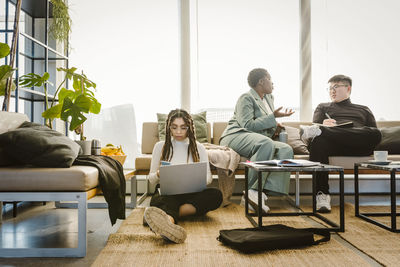 Programmer using mobile phone sitting on carpet while colleague discussing in creative office