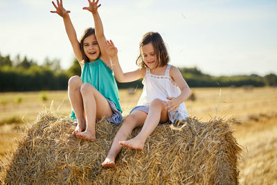 Two charming girls are sitting on a roll of mown rye in a field in summer