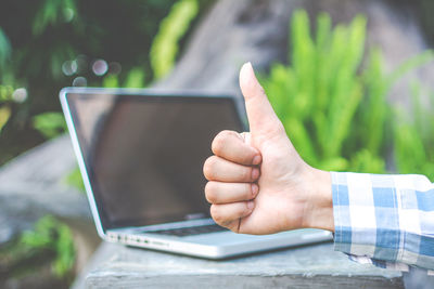 Cropped hand of man gesturing thumbs up sign with laptop in background