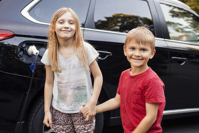 Portrait of happy girl and boy standing against black electric car