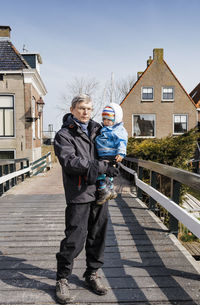 Portrait of grandfather carrying baby girl while standing on footbridge against houses